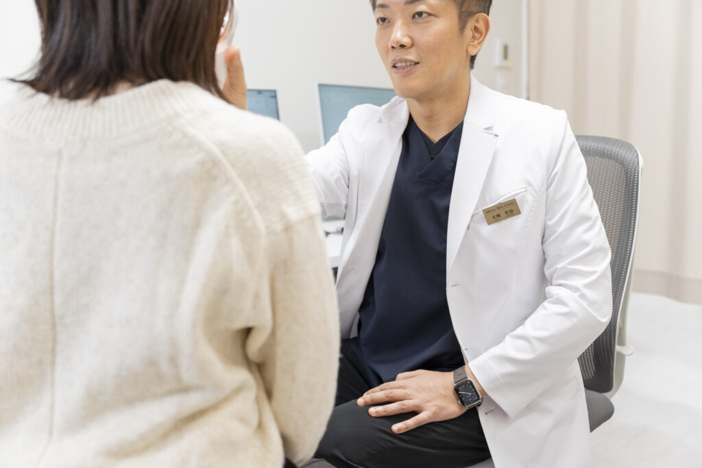 Doctor in a white coat and navy scrubs consults with a patient across a desk in a clinic.